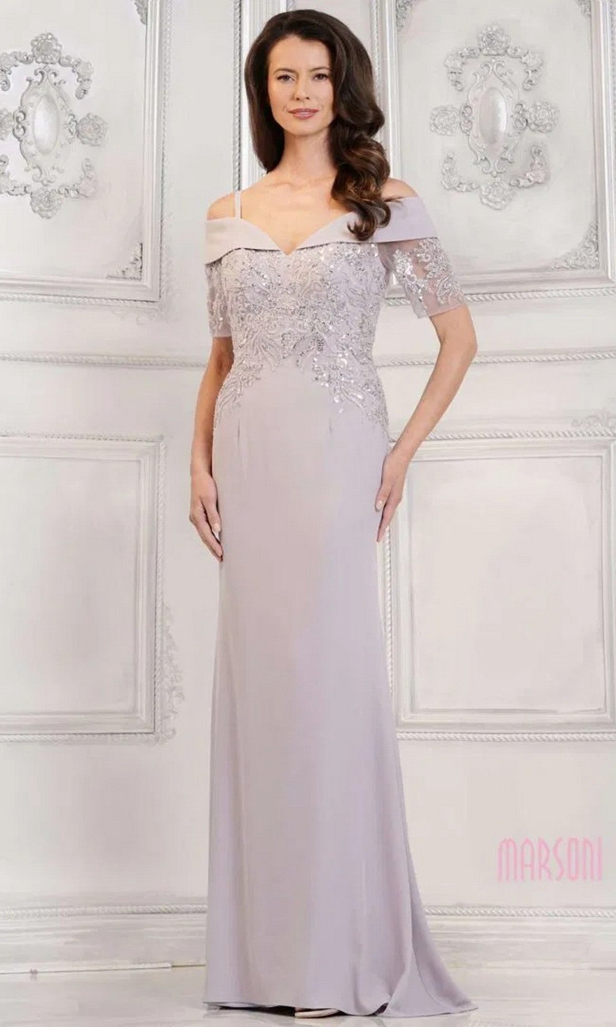 A woman wearing the Long Formal Dress: Marsoni MV1295 in silver, featuring off-the-shoulder crepe fabric and sequin-embroidered appliques, stands against an ornate white wall.
